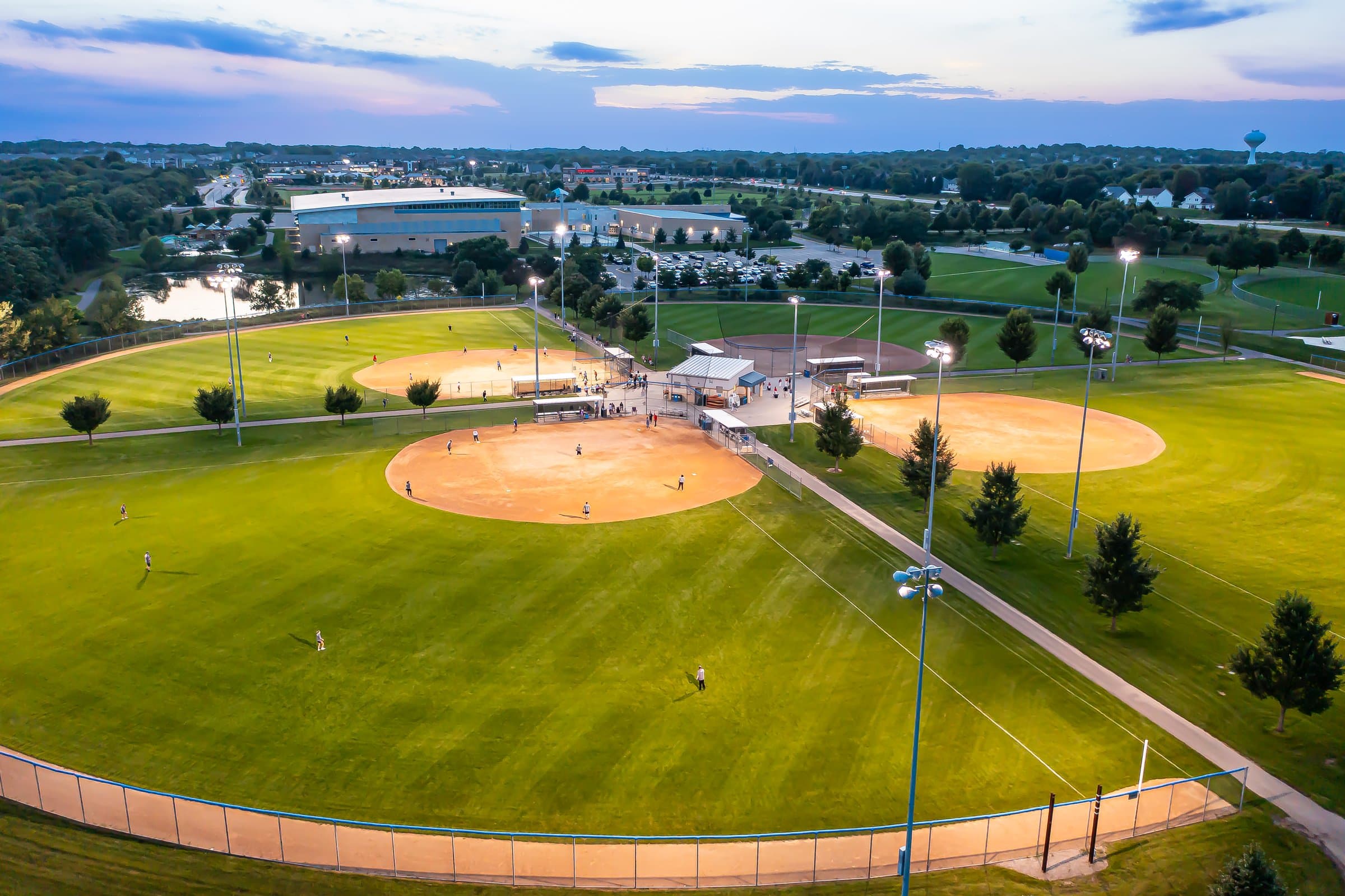 Aerial of Woodbury ballfields at dusk, stadium lights on against a fading sunset sky.